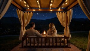 Un couple est assis sur une chaise suspendue, à l’abri d’un auvent, et les étoiles sont visibles dans le ciel nocturne.