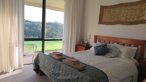 A bedroom with a large bed, a wooden headboard, and a view of a green landscape through a glass door. - Spacious, modern, tranquil, convenient, great location with sea views & bush.  (Tawharanui Peninsula)