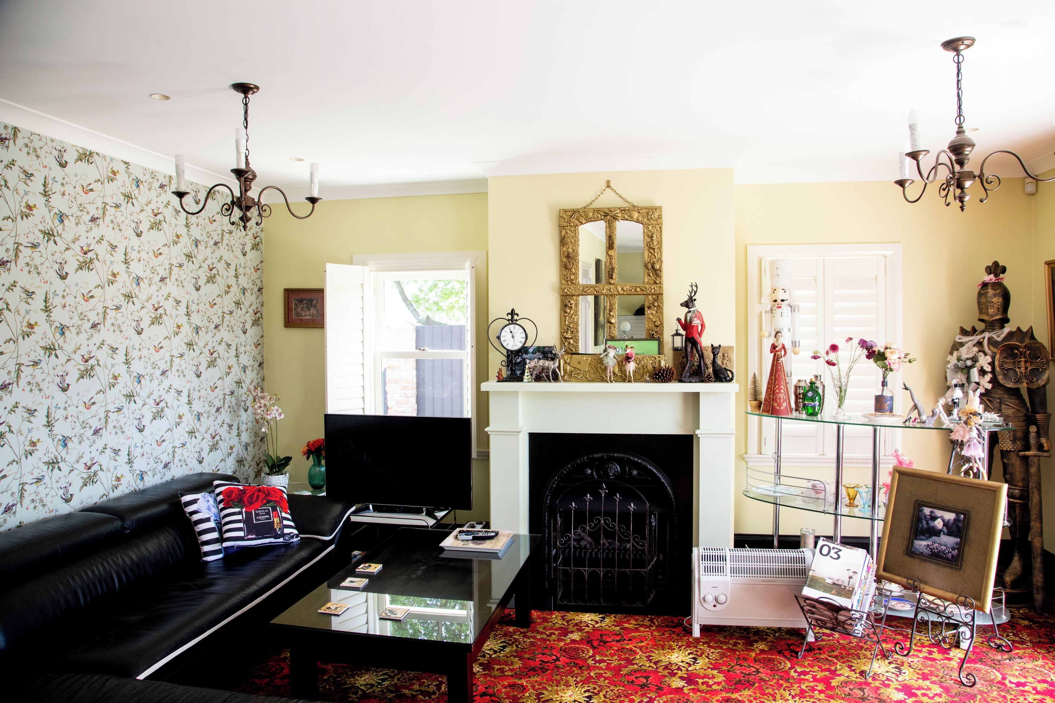 Una sala de estar con un sofá negro, una chimenea, un repisa decorada con adornos y un papel tapiz floral.