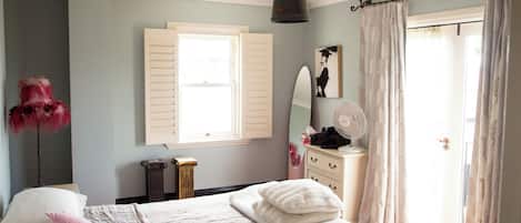 Un dormitorio con cama, cómoda, espejo, radiador, ventana con contraventanas y ventilador.
