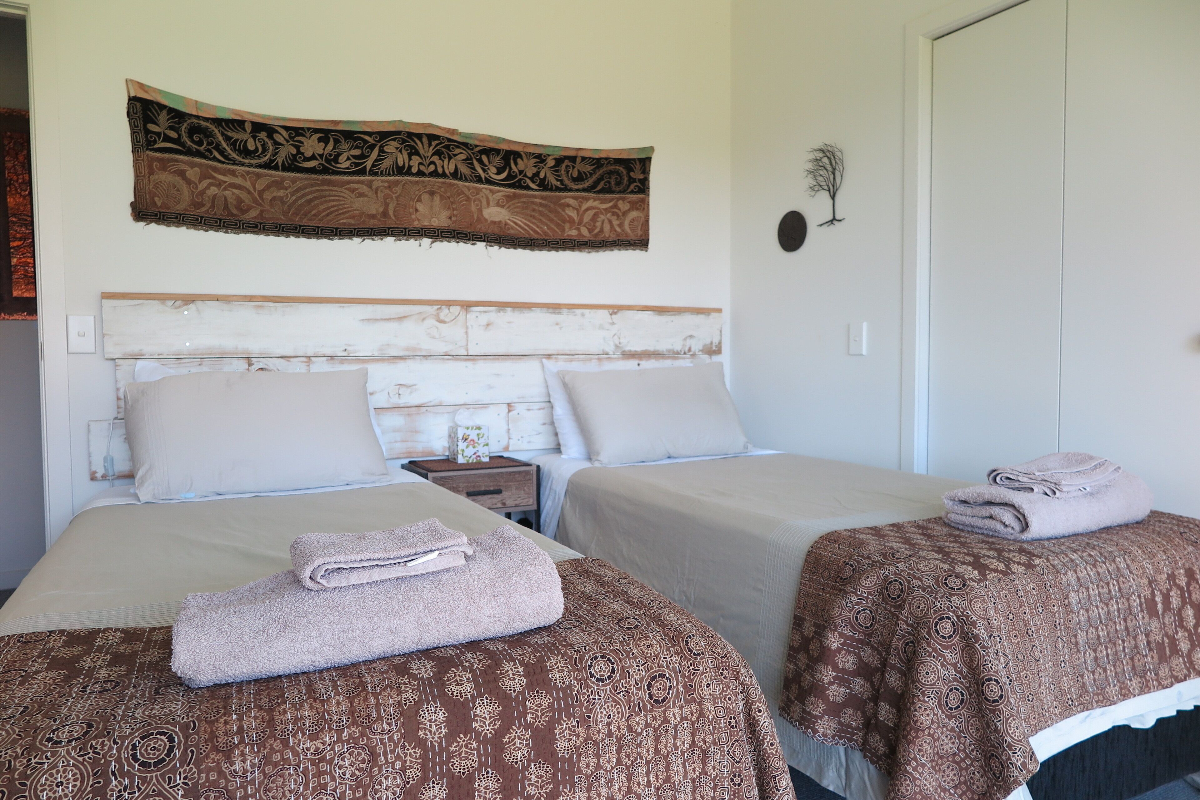 A hotel room with two beds, each with a patterned bedspread and a folded towel. A decorative tapestry hangs above the beds, and a small nightstand with a lamp is visible.