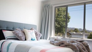 A bedroom with a bed, a grey headboard, a window with curtains, and a view of buildings and trees outside.