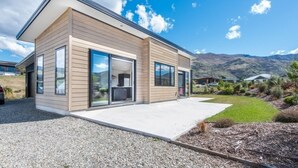 A modern two-story house with large sliding glass doors, a concrete patio, and a mountainous backdrop. - Simply Stunning - Wanaka (FREE WI-FI) (Wanaka)