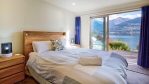 A bedroom with a large bed, a nightstand, a lamp, and a view of a lake and mountains. - 5 Bedroom Queenstown Lakefront Home (Kelvin Heights)