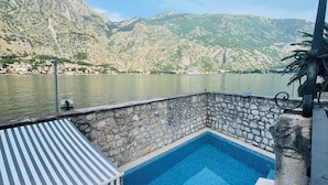 A swimming pool with stone walls, a striped awning, and a view of mountains and a lake.