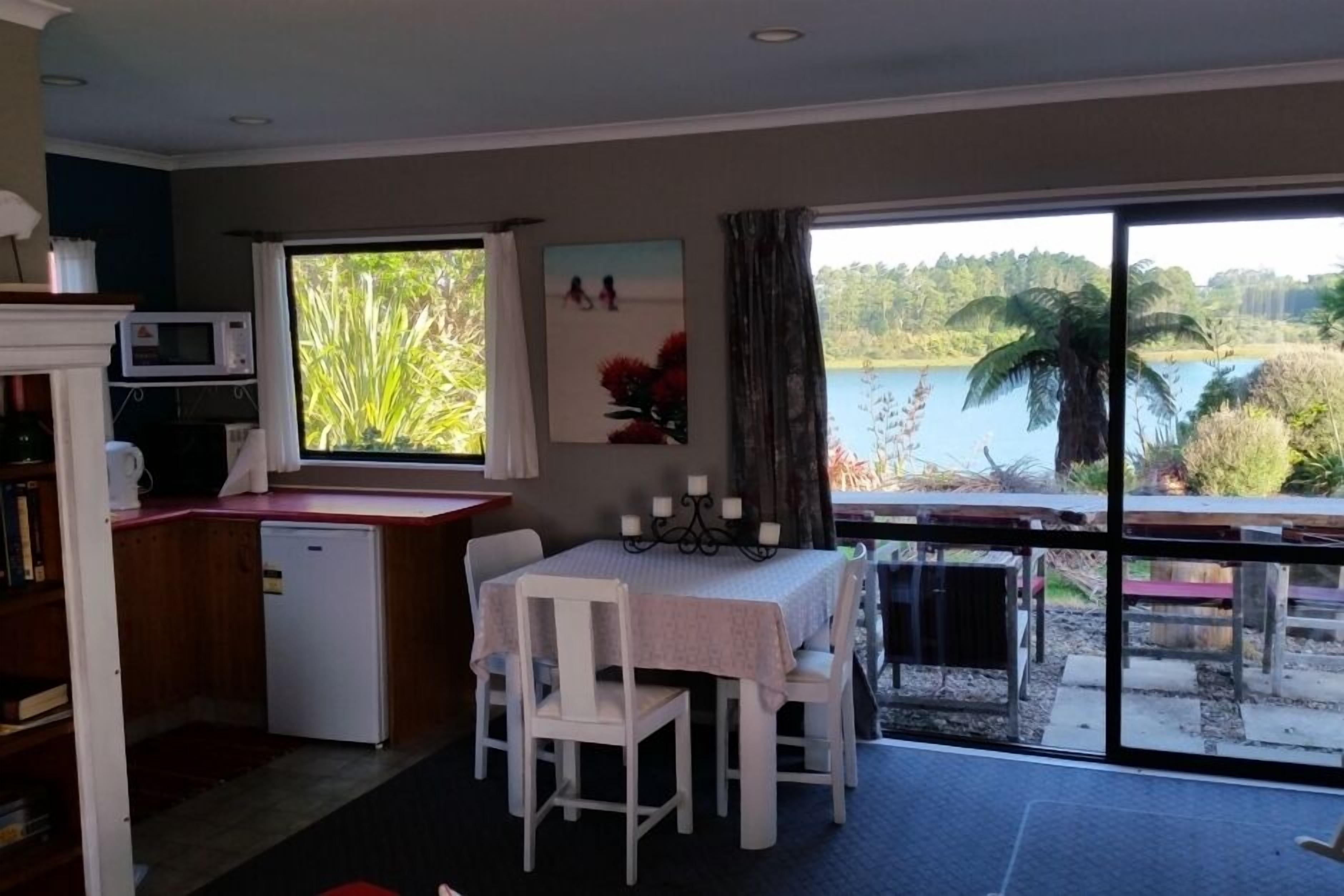 Un comedor con mesa y sillas, una pequeña cocina con microondas, y vista a un lago a través de amplios ventanales.