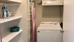 Un cuarto de lavandería con una secadora blanca, estantes con una cesta y un planchador.