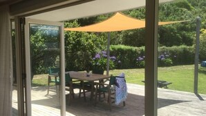 Una puerta corrediza de vidrio abierta que da a un patio exterior con terraza de madera, una mesa con sillas y un toldo amarillo.