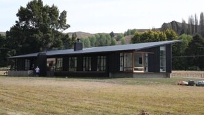 A modern house with a large glass window and a flat roof, surrounded by a grassy field and trees.