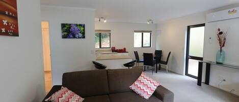 Un salon moderne avec un canapé marron, deux coussins à motifs, un coin repas avec une table et des chaises, et une cuisine en arrière-plan.
