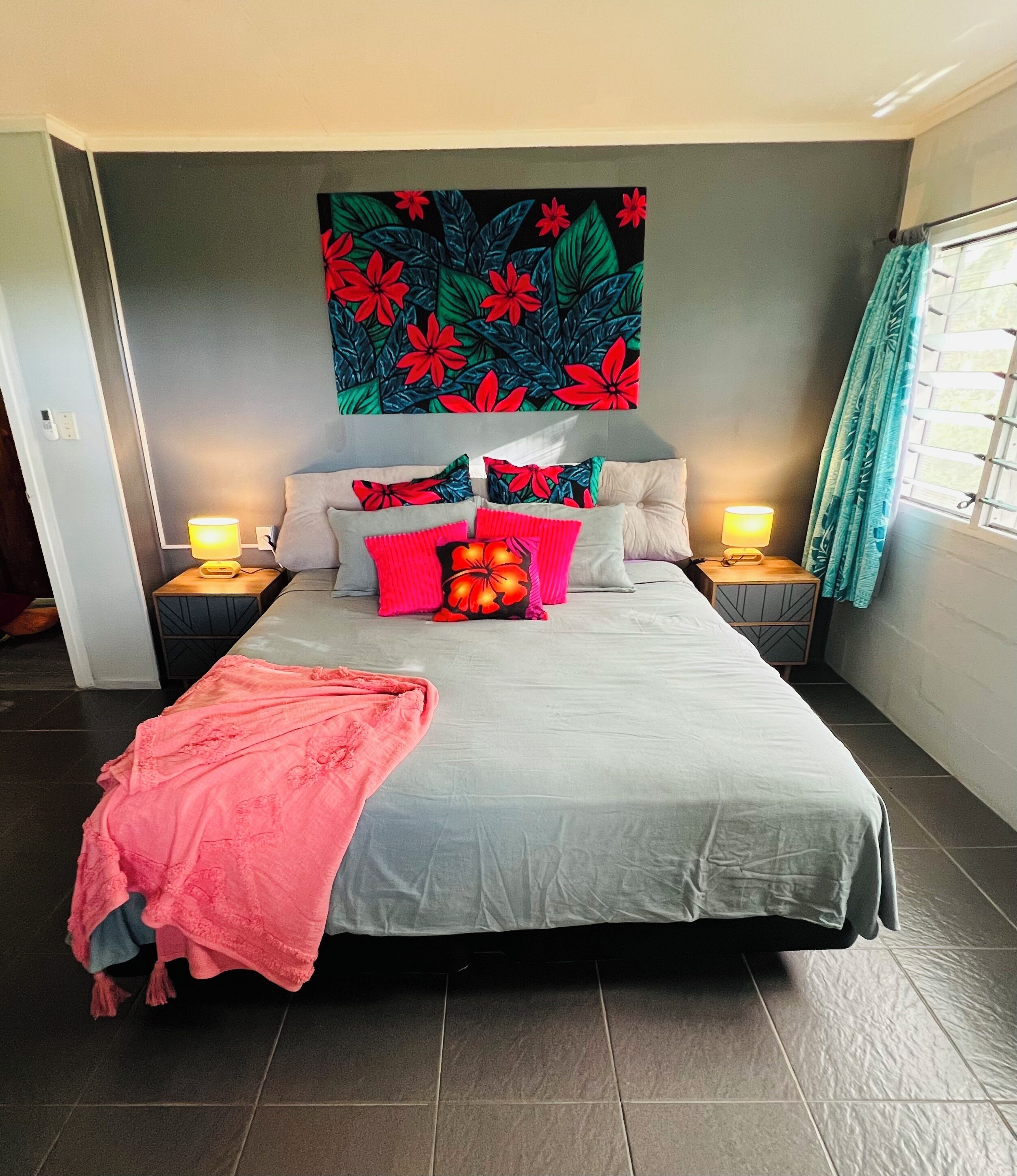 A bedroom with a large bed, two bedside tables with lamps, a floral tapestry on the wall, and a window with curtains.