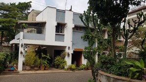 Una casa de dos pisos con balcón, rodeada de abundante vegetación y plantas en macetas.