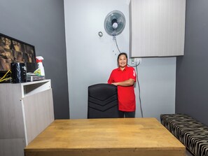 Una habitación con una mesa de madera, una silla de oficina negra, un ventilador de pie y una cama con colcha de estampado de cebra.