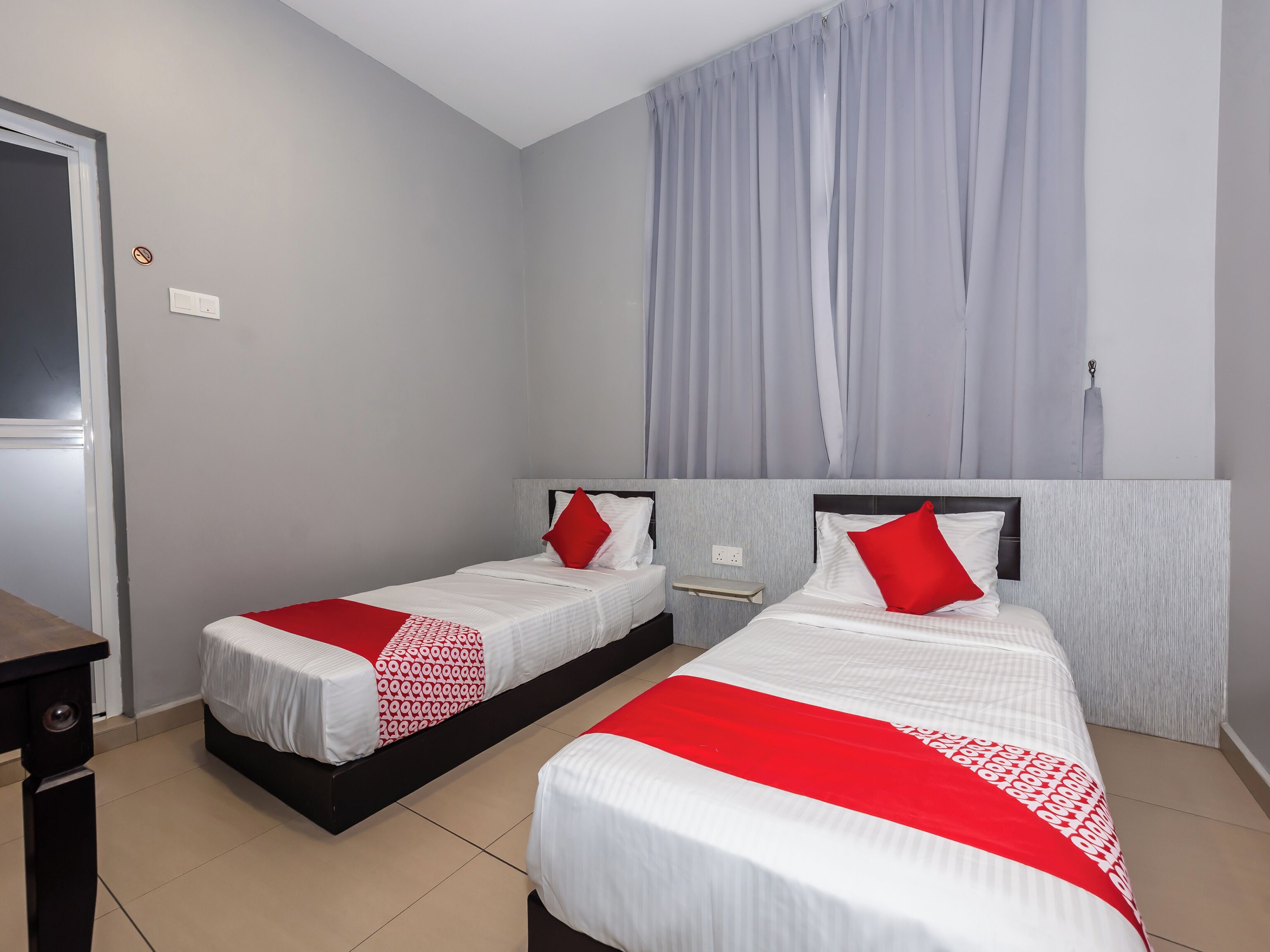A hotel room with two single beds, a desk, a chair, a window with curtains, and a door.