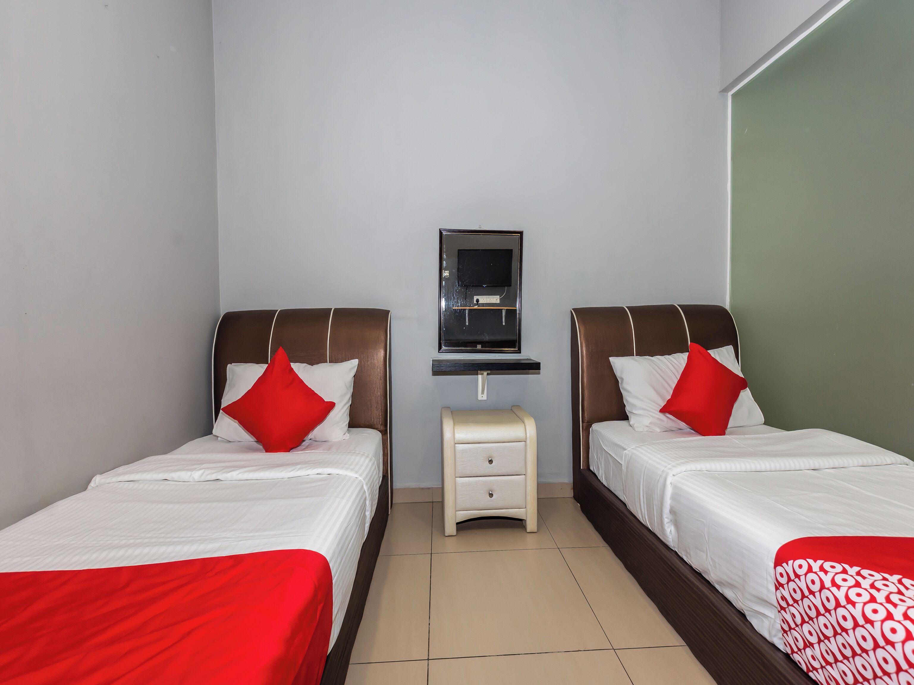 A hotel room with two beds, each with a red pillow, a small bedside table, a TV mounted on the wall, and a green accent wall.