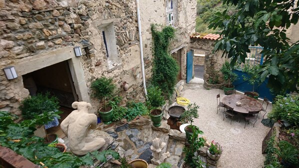 Une cour pavée de pierres, avec un mur en pierre rustique, une petite statue, des plantes en pot, une table avec des chaises et une porte bleue.