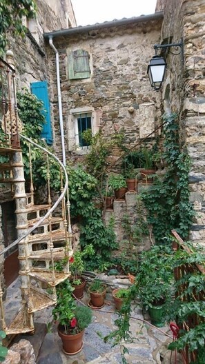 Eine enge Steintreppe mit Blumentöpfen, einem spiralförmigen Metallgeländer und einem rustikalen Steinhaus mit kleinem Fenster und blauem Fensterladen.