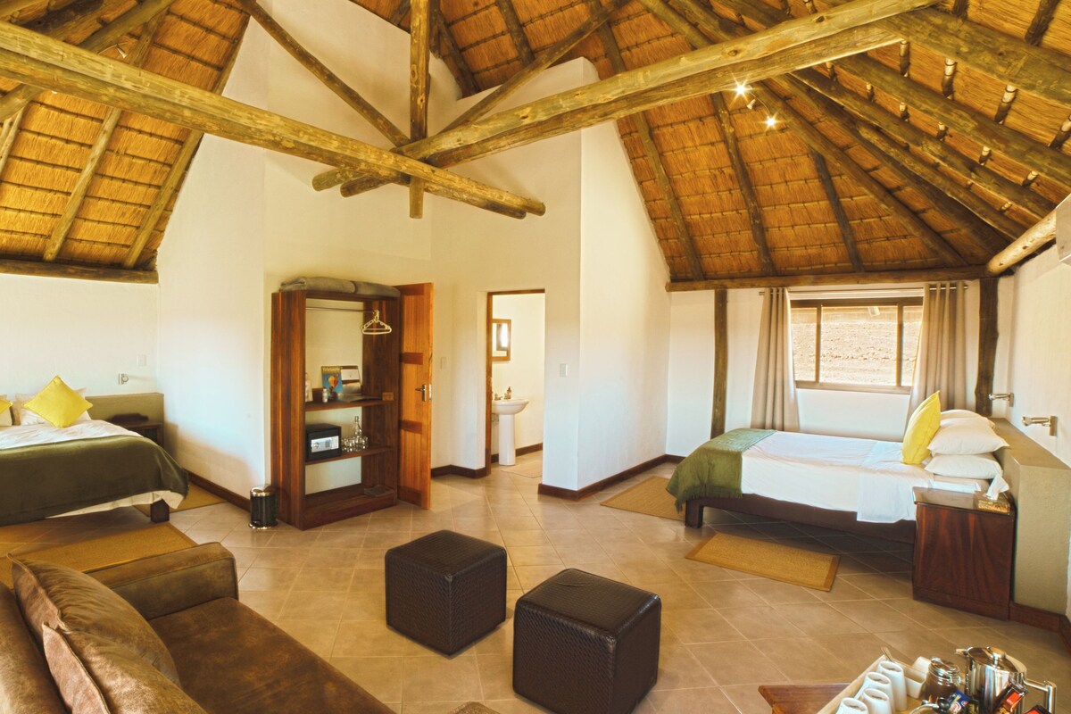 Um quarto espaçoso com teto alto e vigas de madeira, com duas camas com almofadas, um sofá, uma mesinha e um banheiro visível por uma porta aberta.