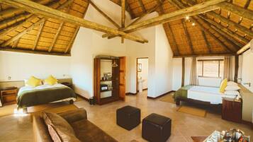 A spacious room with a high ceiling and wooden beams, featuring two beds with pillows, a sofa, a small table, and a bathroom visible through an open door.