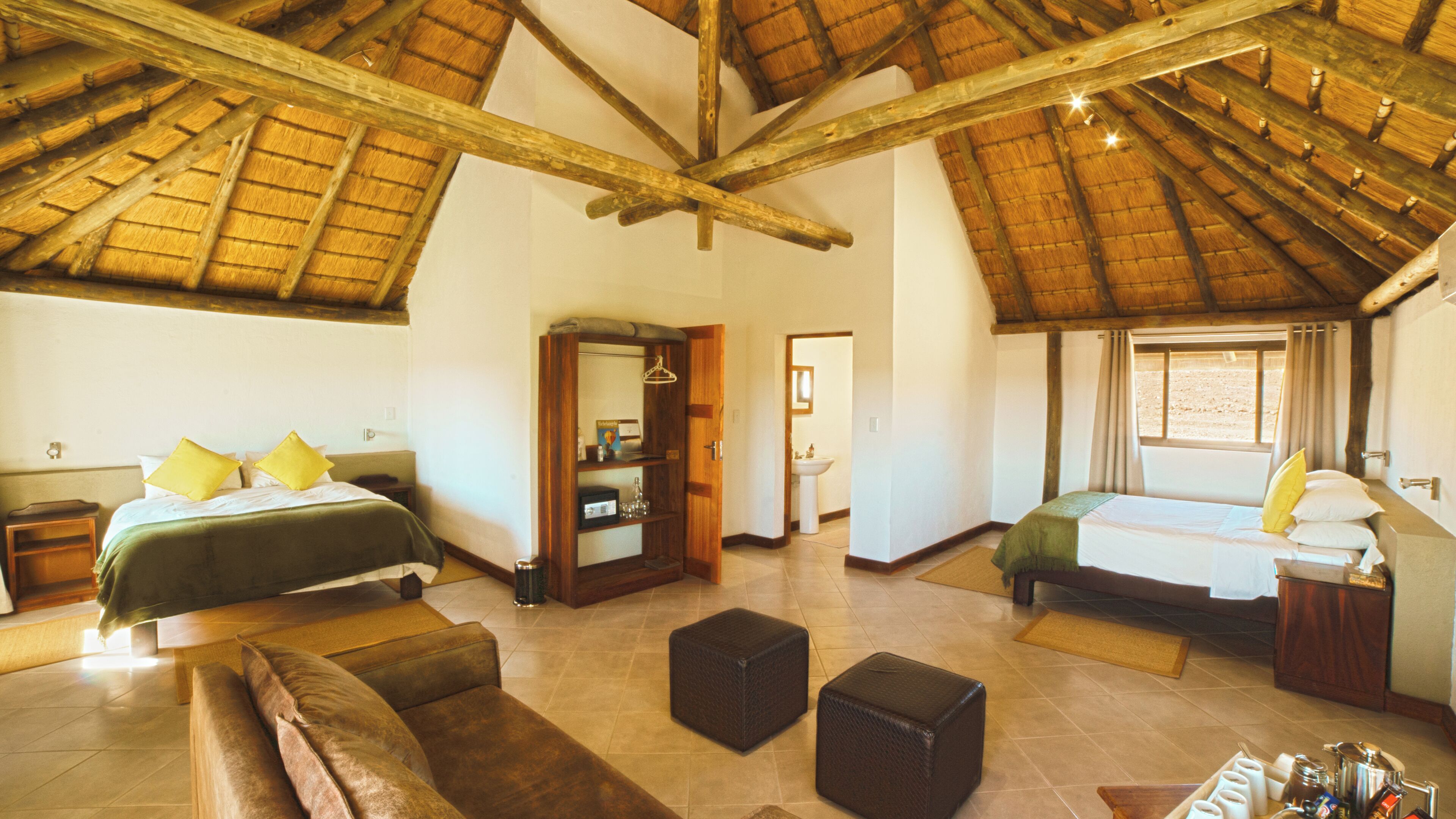 A spacious room with a high ceiling and wooden beams, featuring two beds with pillows, a sofa, a small table, and a bathroom visible through an open door.