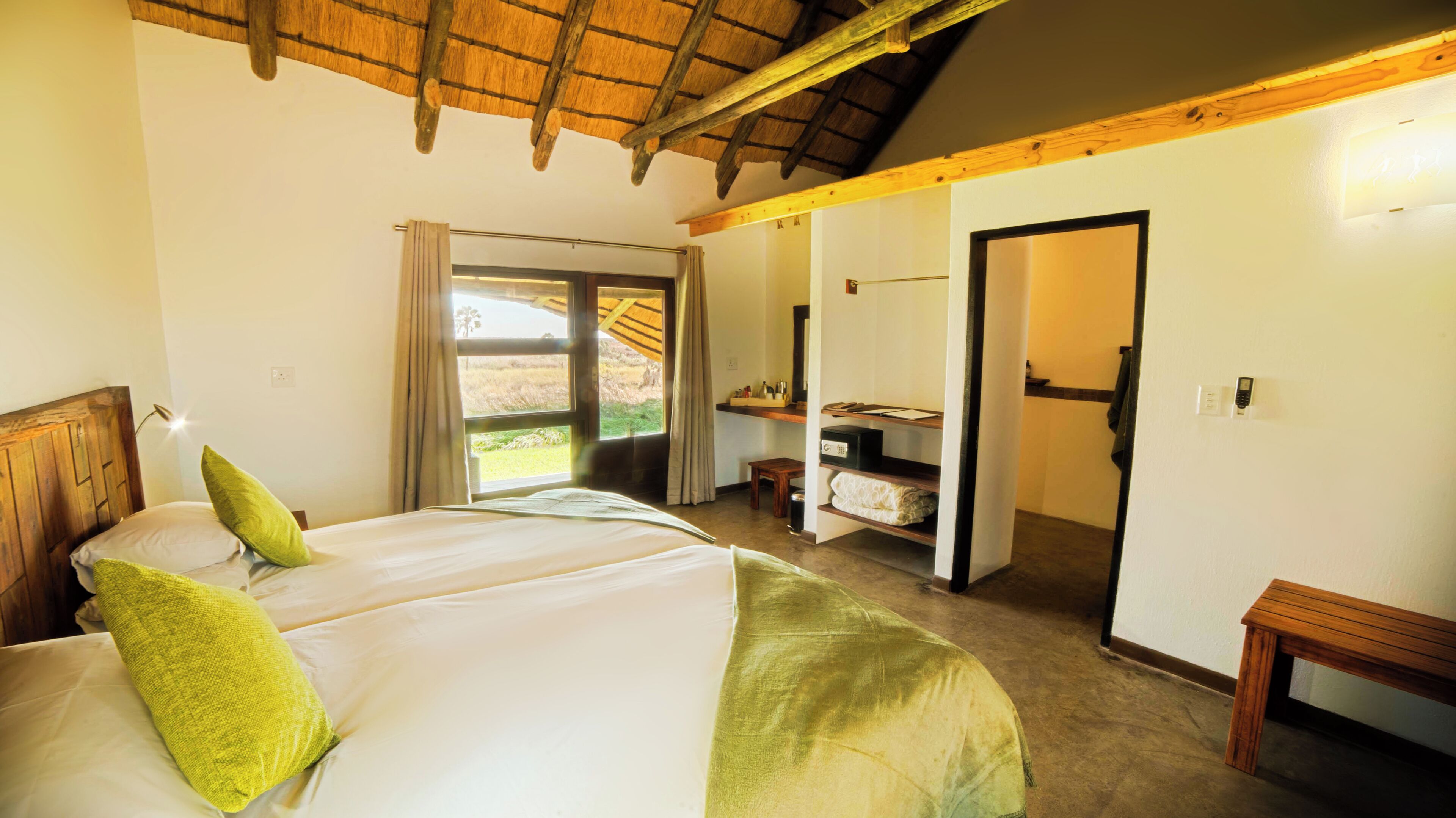 A bedroom with a bed, a nightstand, a window with curtains, a wooden ceiling, and a door leading to another room.