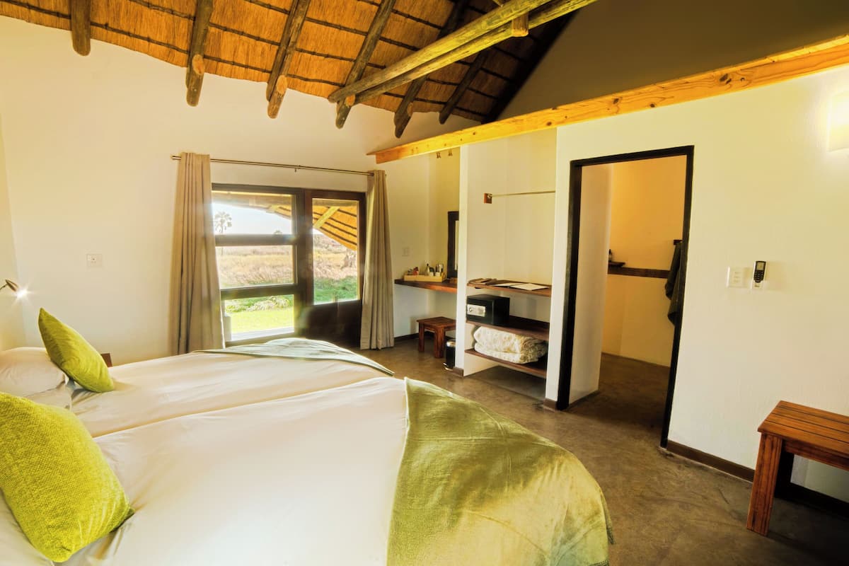 A bedroom with a bed, a nightstand, a window with curtains, a wooden ceiling, and a door leading to another room.