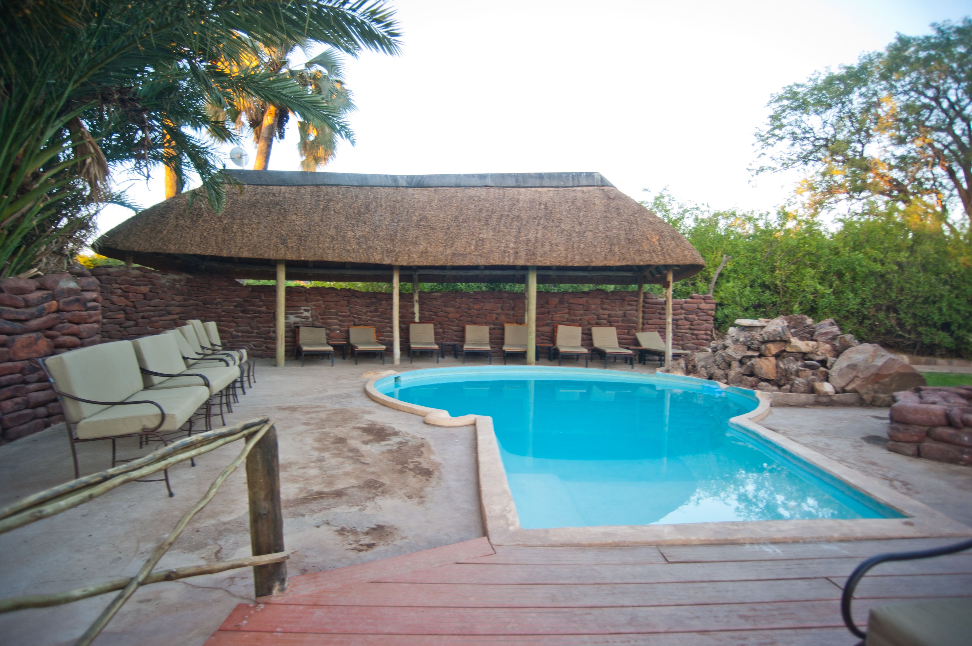 A swimming pool with a thatched roof structure, surrounded by lounge chairs and a stone wall.