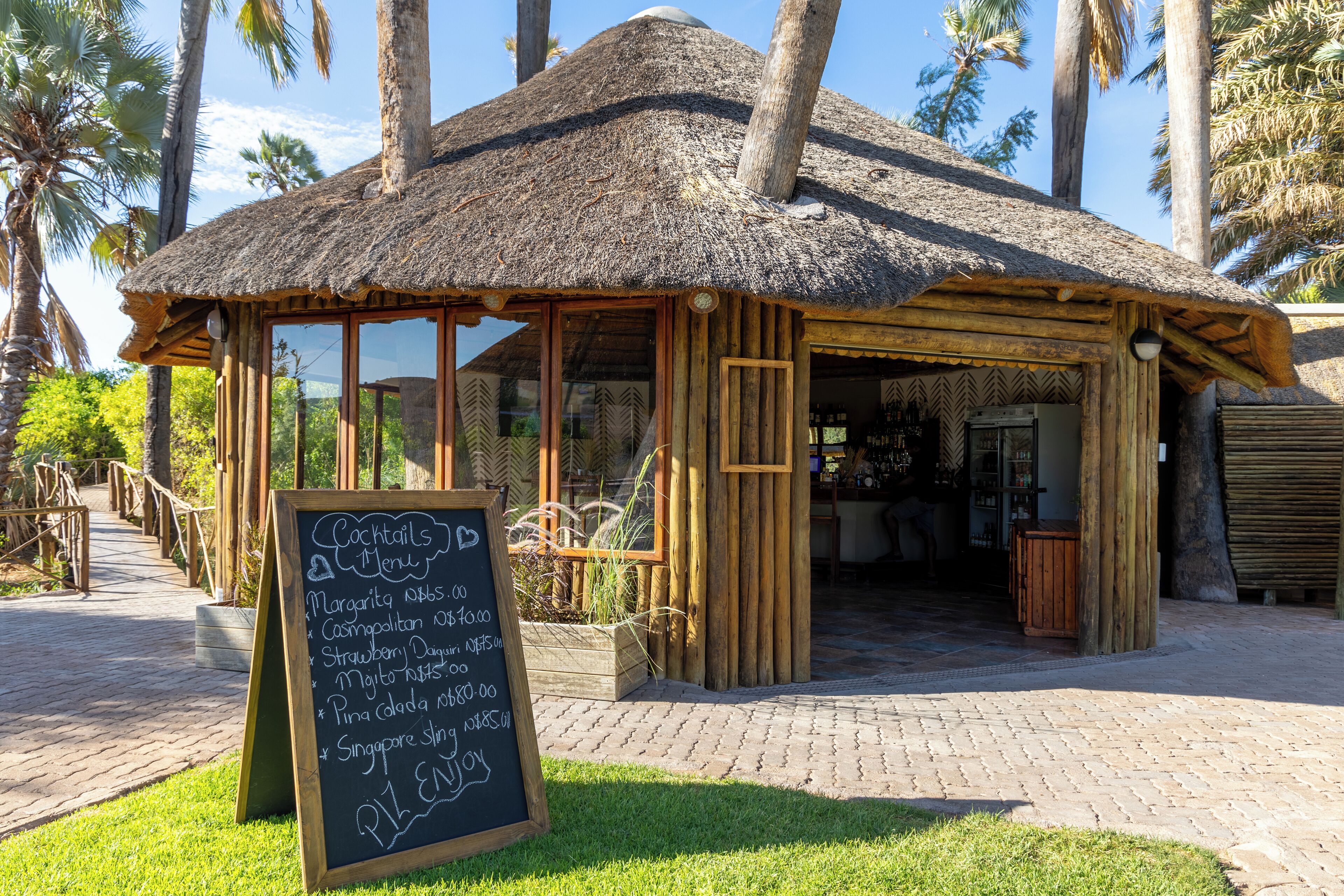 A thatched-roof structure with a wooden facade, a chalkboard menu, and a paved walkway.