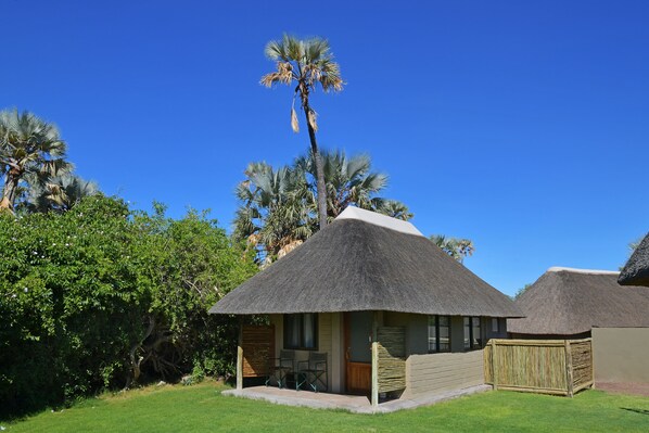A thatched-roof bungalow with a patio, surrounded by lush greenery and palm trees. - Palmwag Lodge (Palmwag)