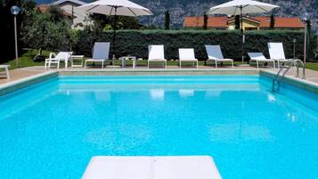 Una piscina cristalina con sillones de baño blancos y sombrillas, rodeada de un jardín ajardinado y un paisaje montañoso.