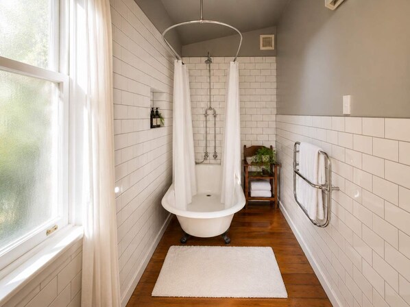 A bathroom with a freestanding bathtub, a shower curtain, a wooden shelf with towels, and a window.