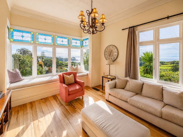A room with a large window seat, a sofa, a red chair, a clock, and a chandelier.