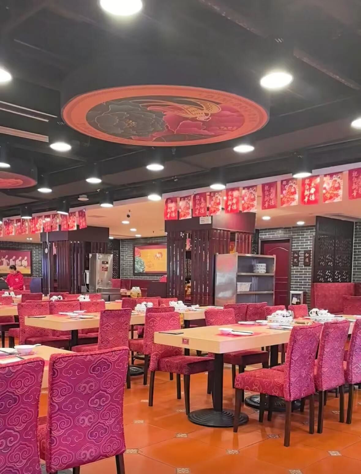 Interior de restaurante com mesas e cadeiras, teto decorativo e faixas vermelhas nas paredes.