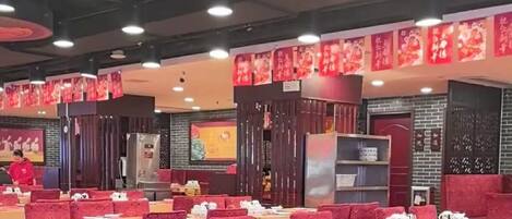 Interior de restaurante com mesas e cadeiras, teto decorativo e faixas vermelhas nas paredes.