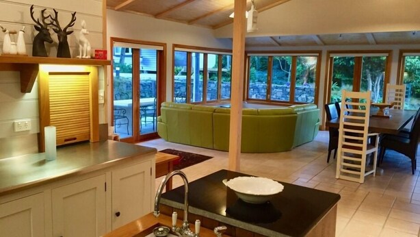 A modern kitchen with a central island, a sink, and a dining area with a green sofa and a wooden table. - Maison du Campagne in French Farm, Akaroa (Wainui (Banks Peninsula))