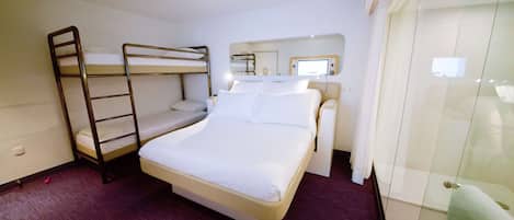 A hotel room with a bunk bed, a single bed, a bedside table with a lamp, a mirror, and a glass shower enclosure.