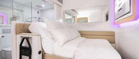 Una habitación de hotel moderna con cama, mesita de noche, espejo y televisor montado en la pared.