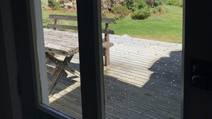 A view through a window with a wooden deck, a stone wall, and a grassy landscape with hills in the distance.