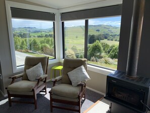 A room with two armchairs facing a large window overlooking a green landscape. A small table and a fireplace are also visible. - A warm, cosy home perfect for your Catlins visit (The Catlins)