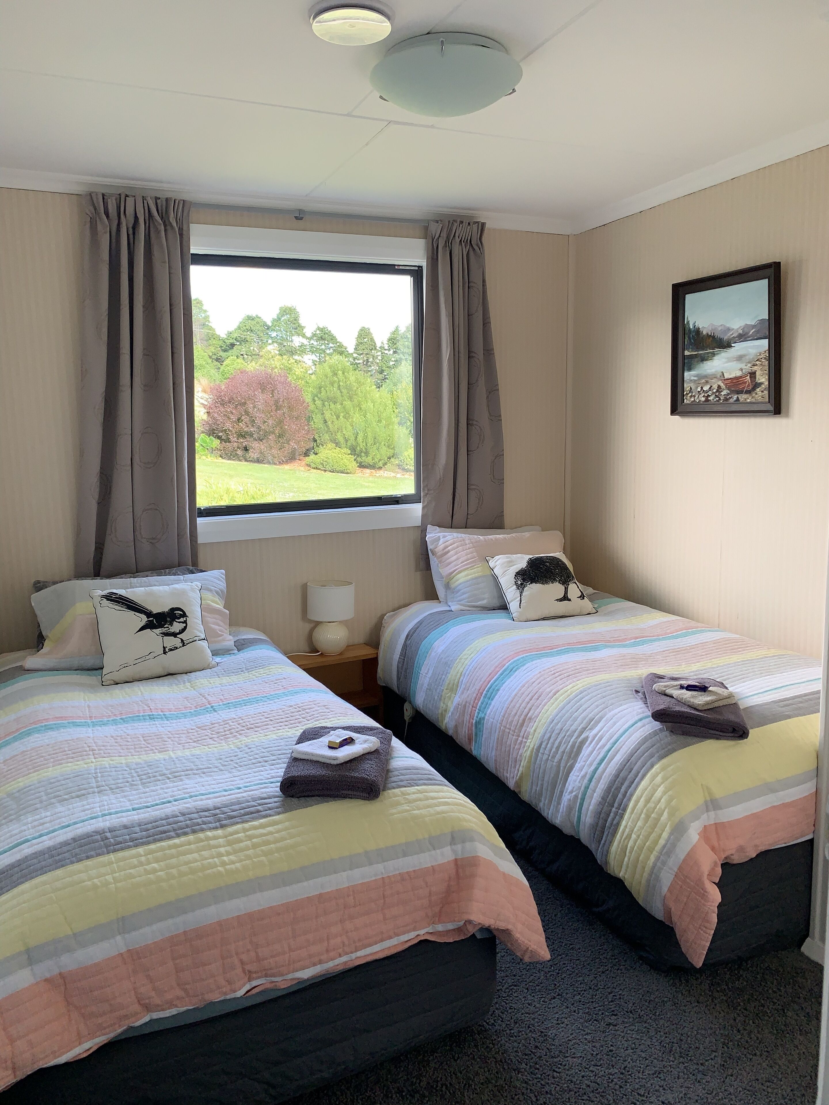 A room with two beds, each with striped bedding and pillows, a window with curtains, a bedside lamp, and a framed picture on the wall.