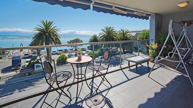 A balcony with a view of the sea, equipped with metal chairs, a small table, and a ladder. - Waterfront Escape - Nelson Waterfront Apartment (Nelson City)
