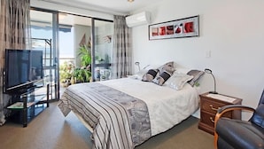 A hotel room with a bed, a TV, a desk, and a chair. There is a balcony with plants and a view of the sea. - Waterfront Escape - Nelson Waterfront Apartment (Nelson City)