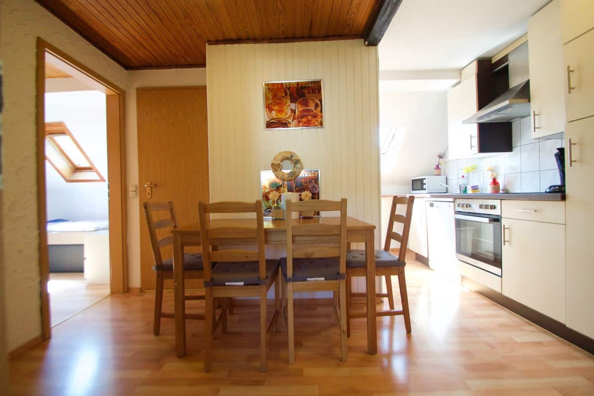 A dining area with a wooden table and chairs, a kitchen with white cabinets and appliances, and a wooden ceiling.