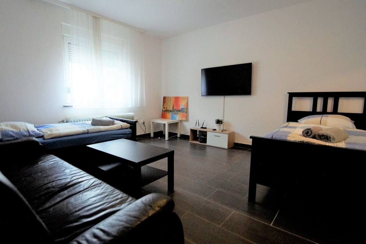 A modern, well-lit room with a black leather sofa, a bed with a headboard, a small white table, a TV mounted on the wall, and a window with blinds.