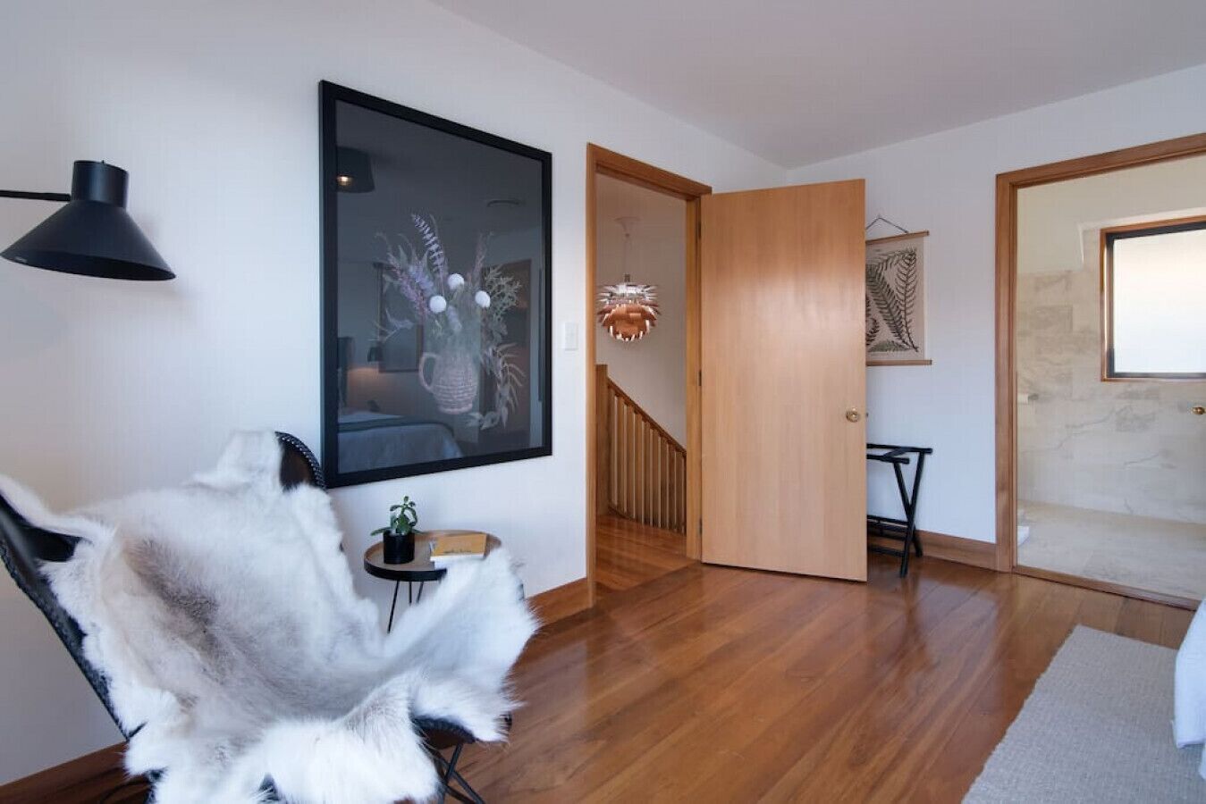 A modern living room with wooden flooring, a black-framed picture, a small round table with a plant, and a white furry chair.