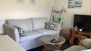Uma sala de estar com um sofá cinza, uma mesa de centro de vidro, uma cadeira de vime, um rack para TV e uma estante com objetos decorativos.