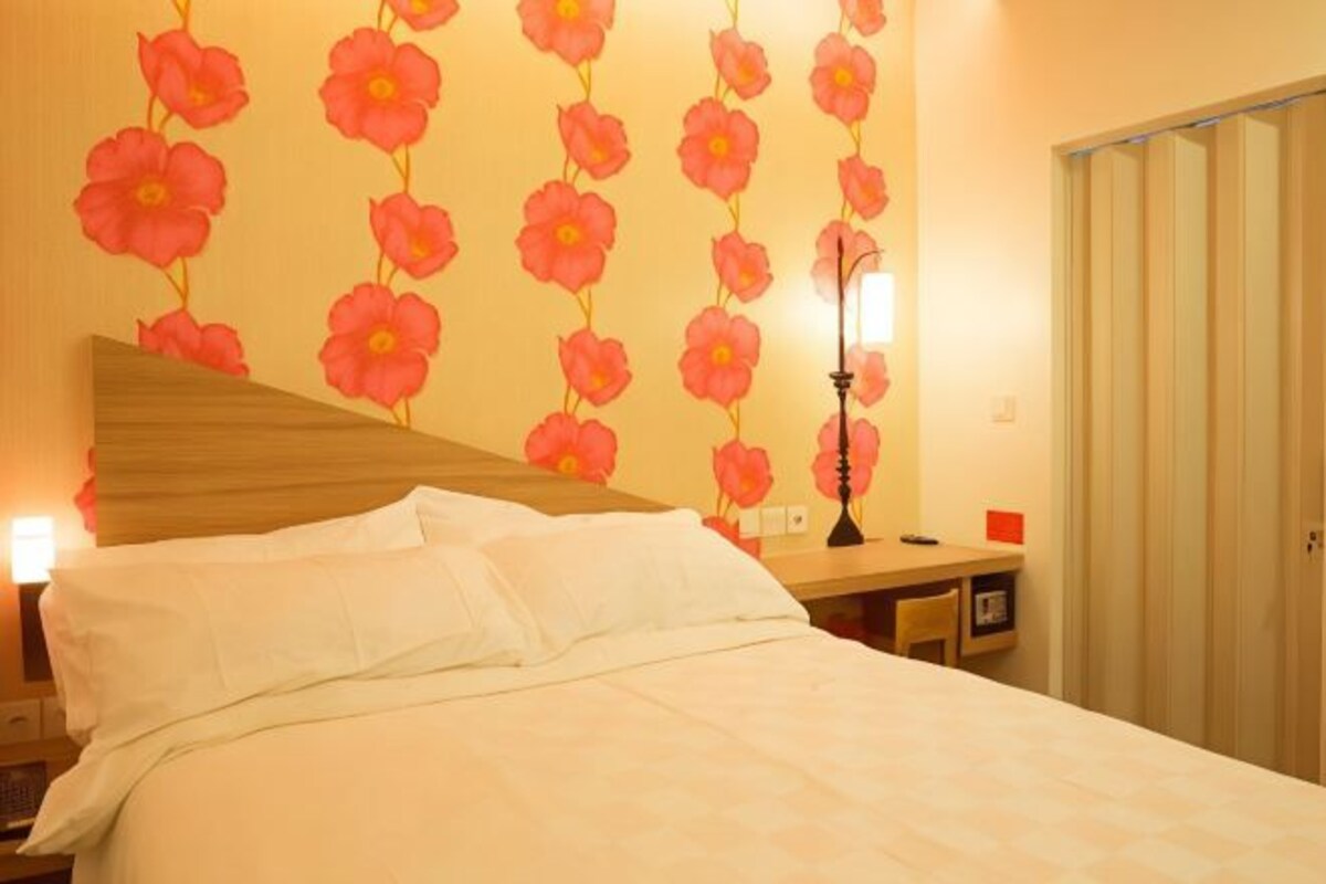 A hotel room with a bed, a nightstand, a desk, and a wall with a floral pattern.