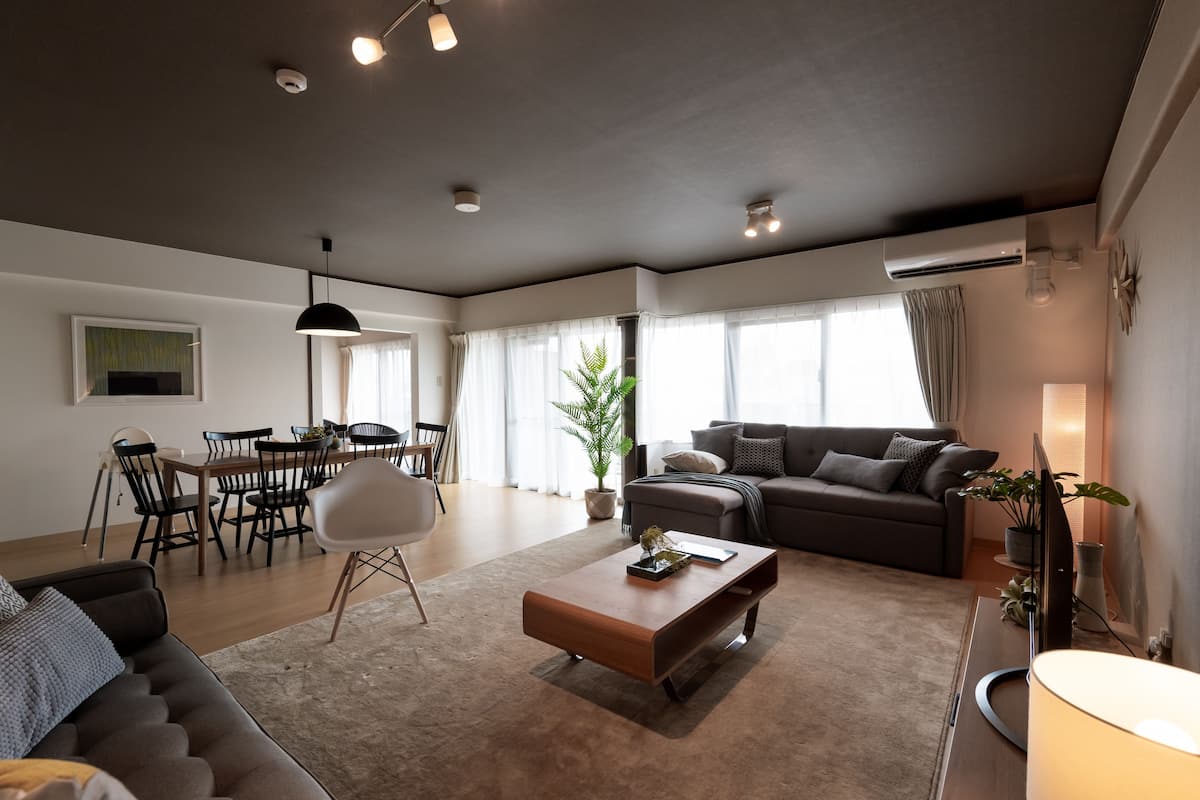 A modern living room with a sofa, a coffee table, a dining area with chairs and a table, and a large window with curtains.