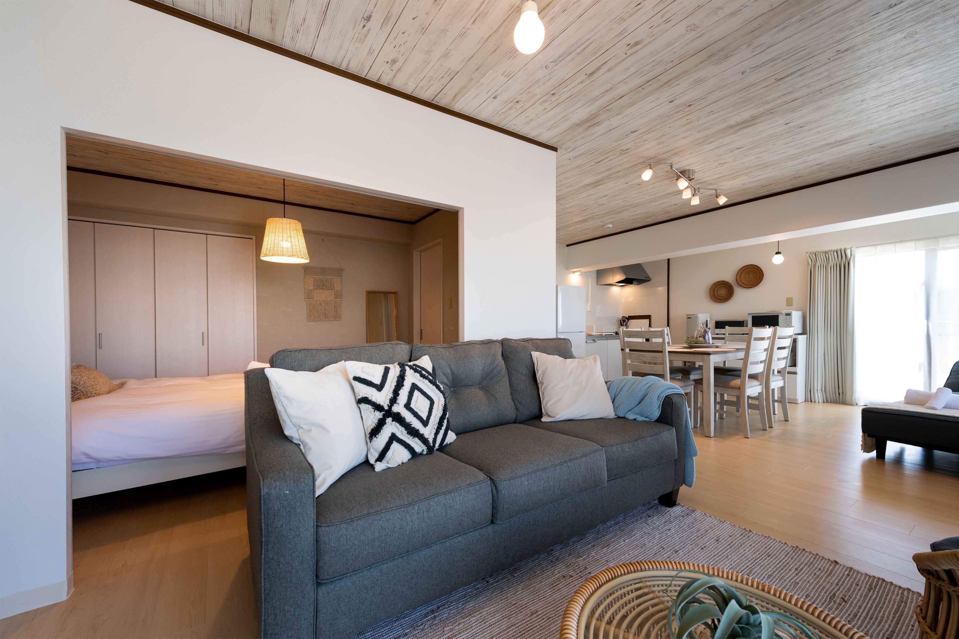 A modern living room with a grey sofa, a bed, and a dining area with a table and chairs.