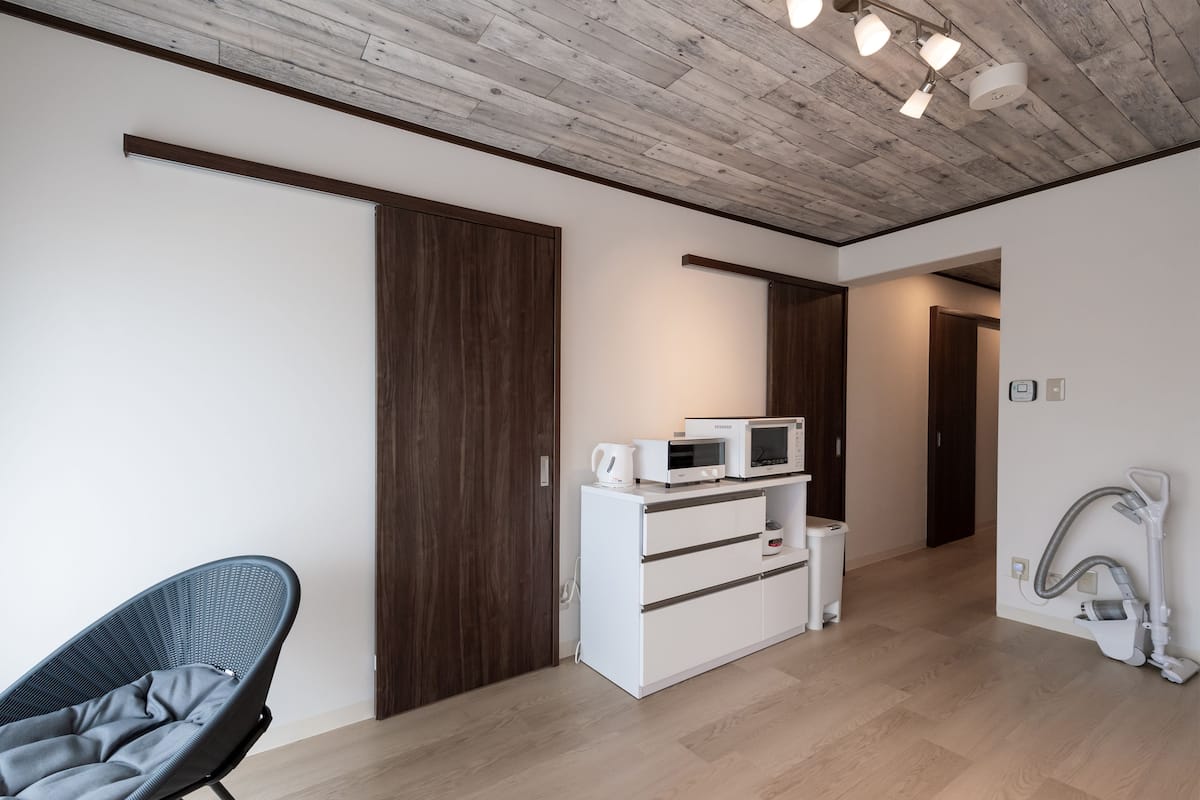 A modern room with a wooden ceiling, a white cabinet with appliances, a grey chair, and a vacuum cleaner.